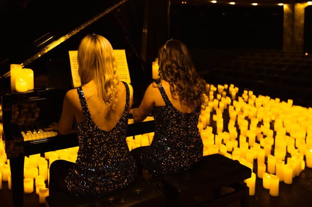 Deux femmes en robes de soirée forme un duo de piano et joue un concert de dos sur un piano à queue noir entourées de bougies comme seule lumière