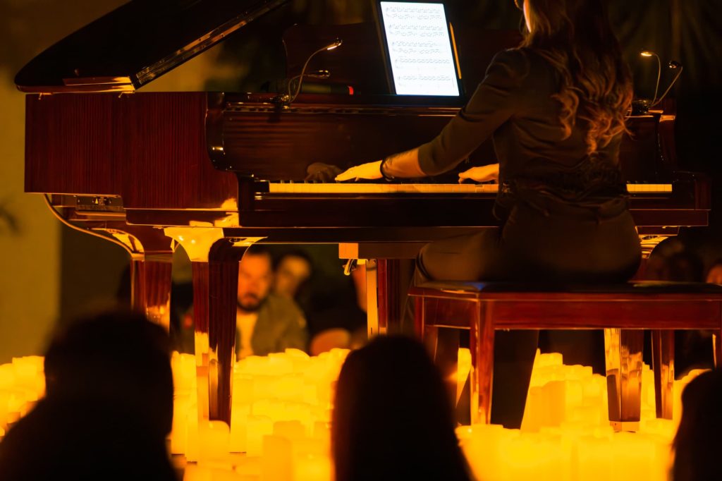 Une pianiste joue un concert sur un piano à queue noir brillant devant un public. Elle est surélevée, est posée sur un sol de bougie qui éclaire l pièce.