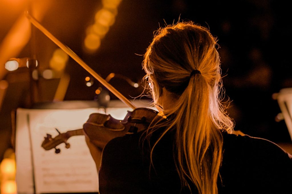 Une femme les cheveux attachés joue un concert au violon de dos en face d'une partition à contre jour, éclairé par une lumière de bougie